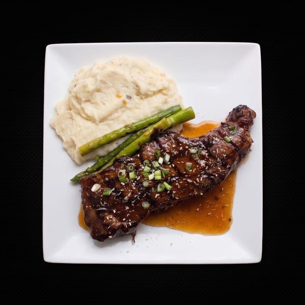 Glazed steak with mashed potatoes and asparagus on a white square plate.