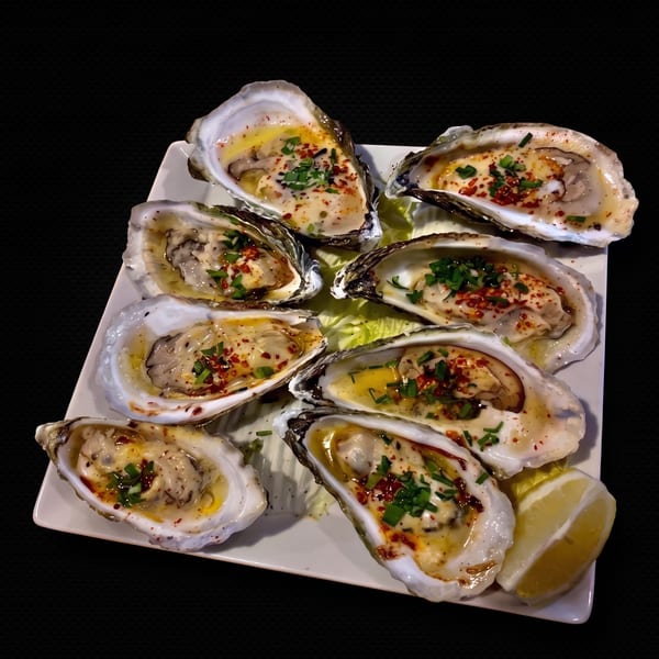 A plate of baked oysters topped with herbs, chili flakes, and a lemon wedge.