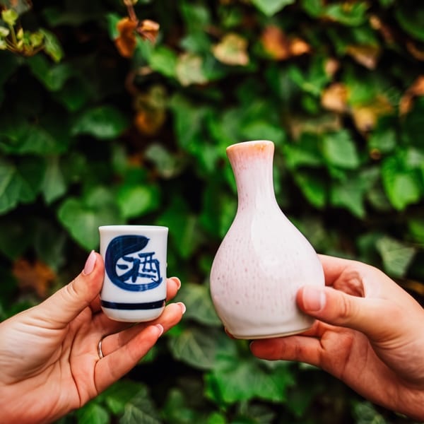 Two hands holding a white sake cup with blue writing and a white sake bottle against green foliage.