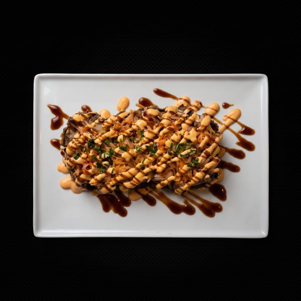 A rectangular white plate holds a dish topped with orange sauce, dark drizzles, and green herbs.