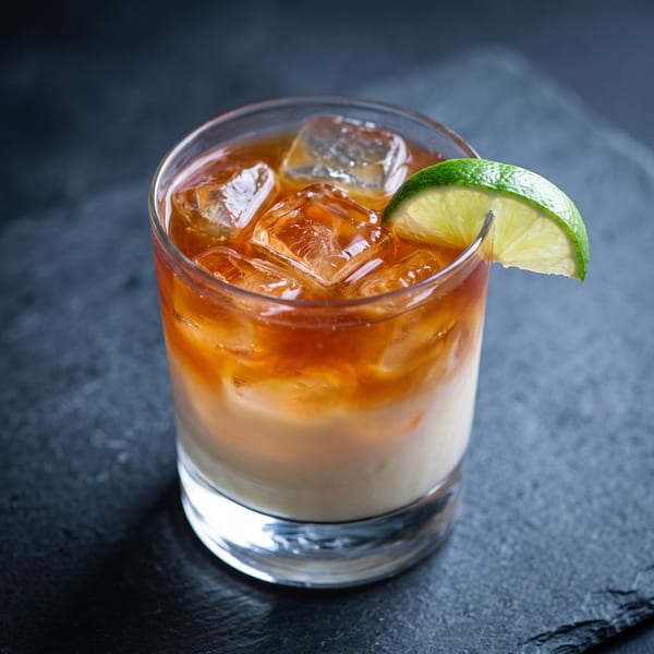 A refreshing cocktail with ice, a dark amber top layer, and a creamy white bottom layer, garnished with a lime wedge.