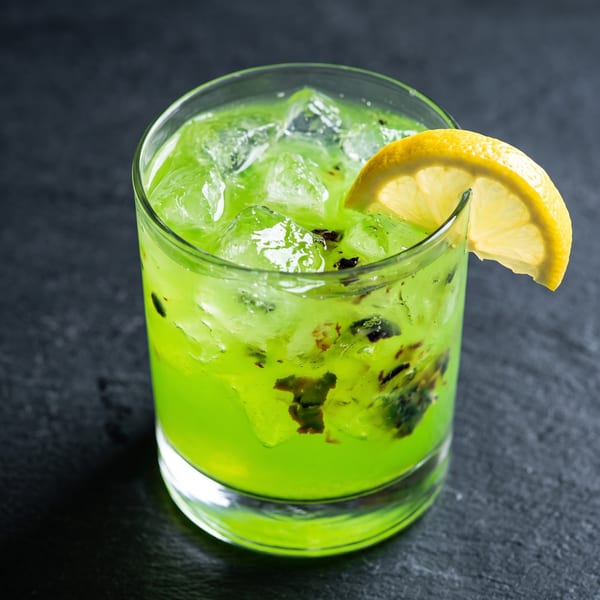 Vibrant green cocktail with ice, kiwi chunks, and a lemon slice on a dark surface.