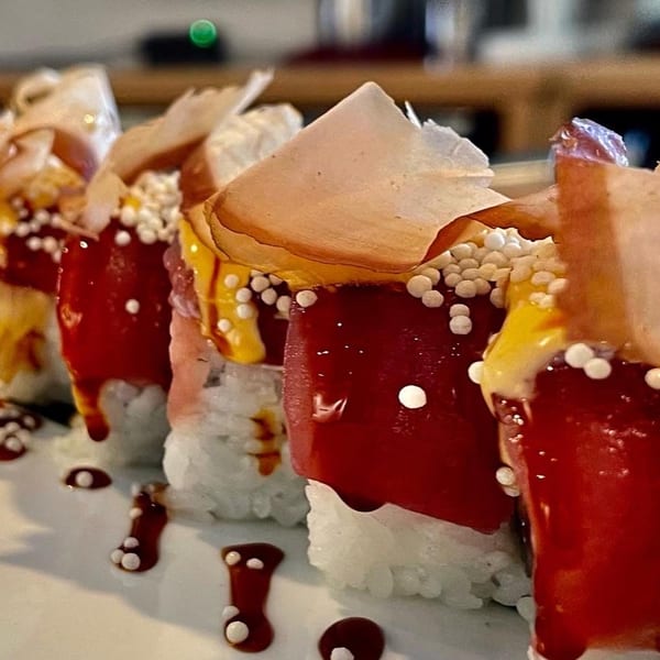 Close-up of sushi rolls topped with red tuna, yellow sauce, white pearls, and bonito flakes.