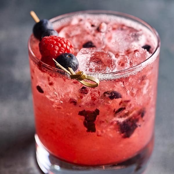 A vibrant red cocktail with ice, garnished with blueberries and a raspberry on a skewer.
